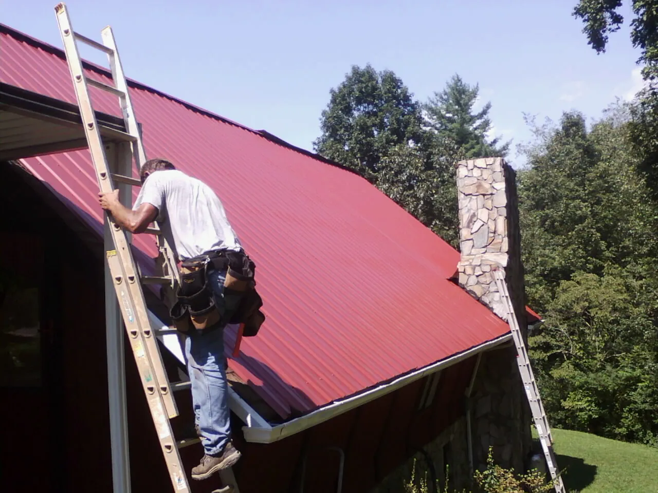 Roofer on a ladder beside a red metal roof and stone chimney — Soffit & Fascia Repair by Roofing Brothers in Montville